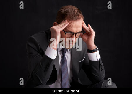 Portraitfotos von einem stressigen im mittleren Alter Geschäftsmann tragen Anzug und dunkler Hintergrund Sitzen mit den Händen auf seine Stirn. Stockfoto