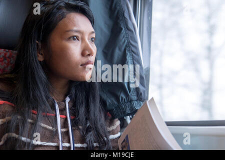 Die junge Frau schaut aus dem Fenster und liest ein Buch in einem Zug. Schöne asiatische Mädchen in Pullover geht in einem Zug mit einem Buch. Stockfoto