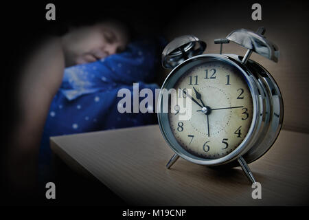 Ein Mann schlafend auf dem Bett neben dem Wecker auf dem Tisch, in der Nacht. Stockfoto