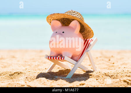 Rosa Sparschwein mit Strohhut gehalten Über den Liegestuhl am Strand Stockfoto