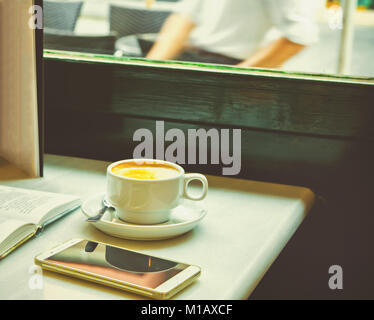 Weiße Tasse mit Cappuccino auf Tabelle mit offenem Buch Smartphone auf Cafe Tabelle durch die Fenster. Holzstuhl, gemütliche Atmosphäre. Im europäischen Stil. Lifestyle Foto Stockfoto