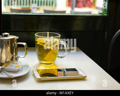 Glas Schale mit frisch Gebrühten dampfend heißen Grüner Tee mit Topf Honig Smartphone am Tisch durch die Fenster in das Cafe. Holzstuhl, Ruhige romantische Gemütliche Atmosph Stockfoto