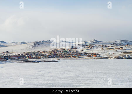 Dorf Landschaft mit Häusern im Winter am Ufer des Baikalsees, die Schiffe auf dem Eis auf einem schneebedeckten Bergen im Hintergrund an einem bewölkten Tag. Stockfoto