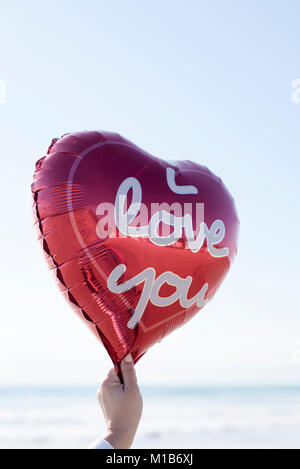Nahaufnahme der Hand einer jungen Frau mit einem roten herzförmigen Luftballon mit dem Text "Ich liebe Dich in Es geschrieben, gegen den Himmel und das Meer in der Bac Stockfoto