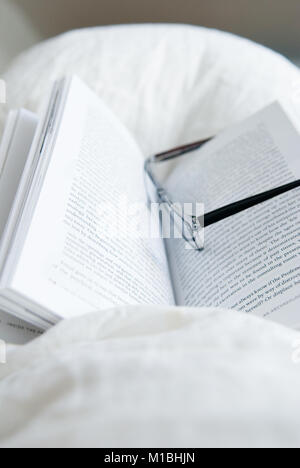 Nahaufnahme der Lesebrille auf einer offenen Taschenbuch auf eine weisse Decke im Bett ausruhen mit Schlafzimmer im Hintergrund Stockfoto