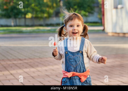 Kleine hübsche Mädchen essen Süßigkeiten im Sommer Park. Adorable kleine Mädchen in Jeans Overalls lächelnd. Porträt eines Kindes 2 Jahre alt, Soft Focus. Stockfoto