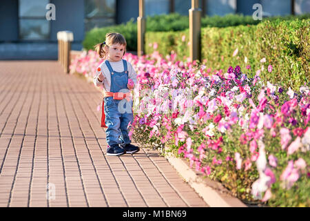 Stilvolle Baby Mädchen 2-3 Jahre alten tragen Jeans Kleidung im Freien. Mit Blick auf die Kamera. Adorable kleine Mädchen in Jeans Overalls. Stockfoto