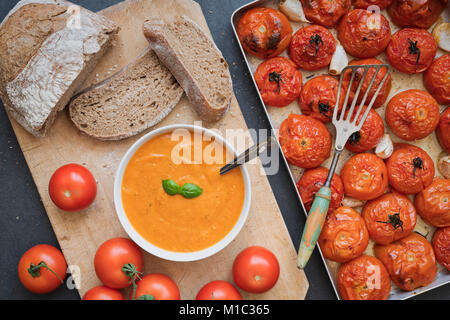 Hausgemachte Tomaten und Basilikum Suppe mit Schwarzbrot Neben gerösteten Tomaten und Knoblauch in einem Backblech auf einer Schiefertafel Hintergrund. Stockfoto