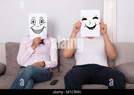 Paar hält lächelnde Gesichter vor den Gesichtern Sitzen auf der Couch Stockfoto
