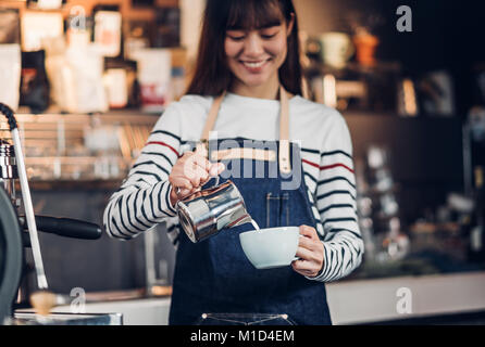 Asien Frau barista gießen Sie Milch in die heiße Tasse Kaffee an der Bar vor der Maschine im Cafe Restaurant, Essen Geschäftsinhaber Konzept Stockfoto