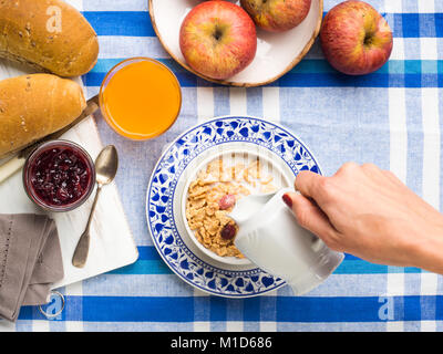 Home Frühstück mit Müsli, Milch, Obst und Brot serviert auf blaue und weiße Tischdecke. Woman's Hand gießen Milch Stockfoto