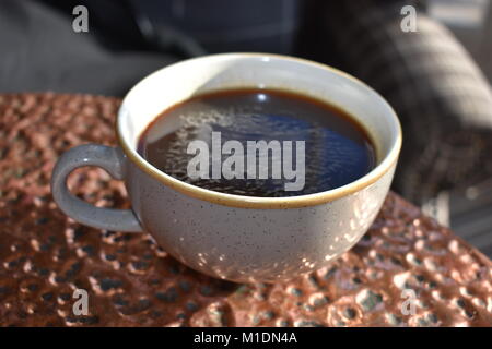 Große graue Tasse schwarzen Kaffee auf Kupfer Tabelle mit einem unscharf Sessel im Hintergrund. Stockfoto