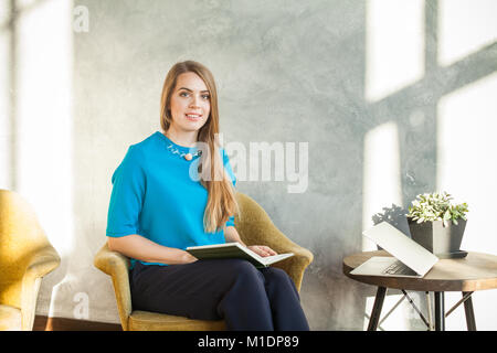 Lächelnde Frau Sitzen im Home Office und Kamera. Attraktives Modell mit Buch und Laptop. Frau Arbeiten mit neuen Startprojekt in modernen Lo Stockfoto