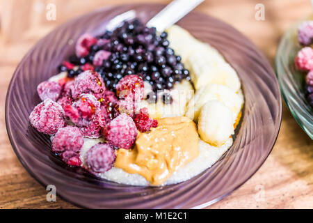 Makro Nahaufnahme von haferflocken Hafer Korn porridge Bowl mit in Scheiben geschnittenen Bananen, Erdnussbutter, gefrorene Beeren, Himbeeren, Heidelbeeren, Ahornsirup Stockfoto