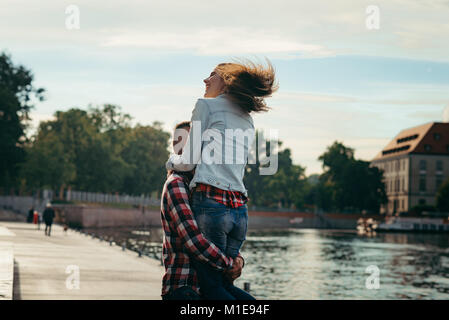 Der junge Mann ist Anheben der fröhliche hübsche Mädchen, das auf dem Hintergrund der Fluss in Polen zu lachen. Ansicht von hinten. Stockfoto