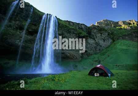 Island. Seljalandsfoss. Liegende Frau in Zelt lesen Buch, der Wasserfall Seljalandsfoss im Hintergrund. Camping Stockfoto