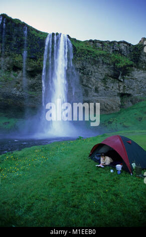 Island. Seljalandsfoss. Liegende Frau in Zelt lesen Buch, der Wasserfall Seljalandsfoss im Hintergrund. Camping Stockfoto