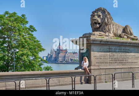 Blonde weibliche Touristische sitzt auf der Attika der berühmten Kettenbrücke in Budapest mit Blick auf das Parlament Gebäude über den Fluss Stockfoto