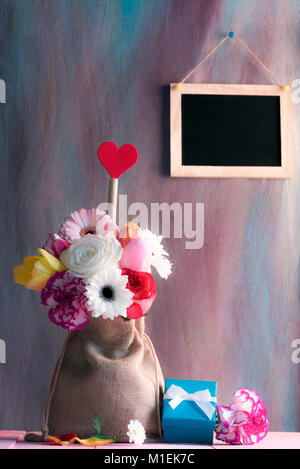 Muttertag Karte mit bunten Blumen und ein rotes Herz Form in einem hessischen Beutel, einem blauen Geschenkbox auf einem rosa Tabelle und eine Tafel auf der violetten Wand. Stockfoto