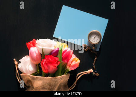 Bunten Strauß unterschiedlichster Blumen in einer Jute-tasche, eine alte Taschenuhr und eine leere blau Nachricht Karte, auf einem schwarzen Hintergrund. Stockfoto