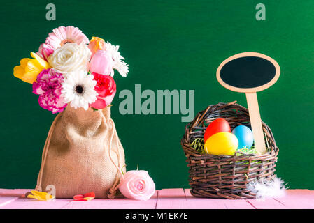 Ostern Karte mit einem Blumenstrauß in einer jutetasche und einen Weidenkorb mit bemalten Eiern und einem hölzernen Banner, auf einer rosa Tisch und einer grünen Wand. Stockfoto