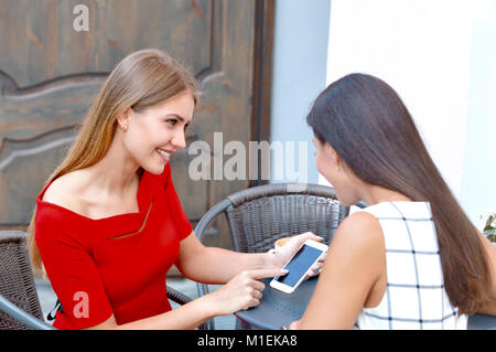 Zwei schöne Frauen trinken Kaffee an der Bar im Freien. Frau zeigt auf einem Handy etwas zu seiner Freundin. Lifestyle und Freundschaft Konzept Stockfoto