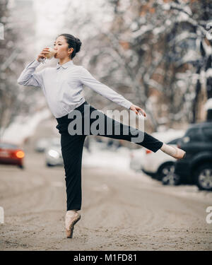 Ballerina in schwarze Hosen und weiße Bluse mit Glas in den Händen gekleidet ist trinken heißen Kaffee und stehen vor Ort Straße gegen Schnee und Winter b Stockfoto