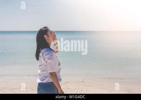 Frau tragen weiße T-Shirt, Sie auf Sand Strand und Holding Sonnenbrille in der Hand mit wunderschönen Blick auf das Meer im Hintergrund. Stockfoto