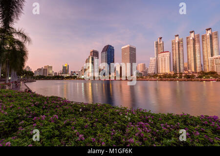 Malerischer Blick auf Blumenbeete, Holzsteg und See an der Benjakiti (benjakitti) Park und Wolkenkratzer in Bangkok, Thailand, bei Sonnenuntergang. Stockfoto