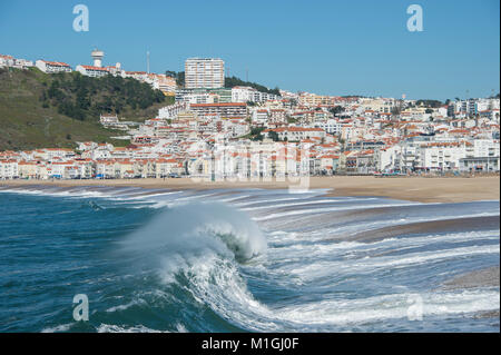Welle bricht auf den Strand von Nazare, Portugal mit der Stadt im Hintergrund Stockfoto