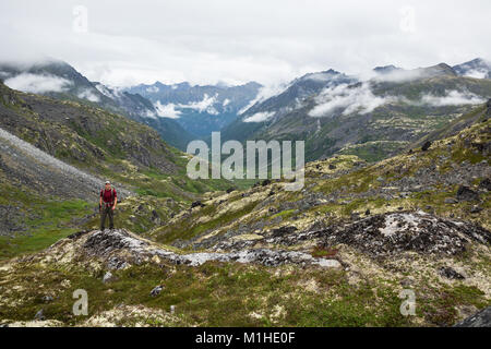 Ein junger Mann steht mit seinem Rucksack und Wanderstöcken auf einem felsigen Aussichtspunkt mit Blick auf eine Glacial River Valley in der Hatcher Pass Bereich von Alaska Stockfoto