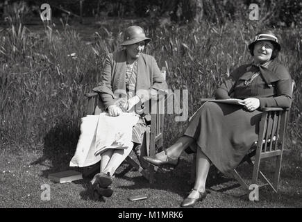 1930er Jahre, historische, zwei englische Damen draußen sitzen in Holz- Leinwand Garten Stühle auf einem Rasen in einem grossen Garten entspannen mit ihren Zeitungen und Buch, England, UK. Stockfoto