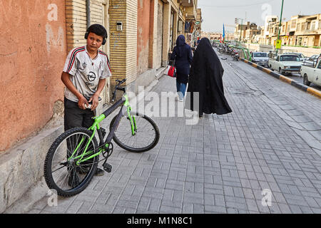 Isfahan, Iran - 24. April 2017: Eine iranische Junge mit einem Fahrrad steht auf der Straße, auf dem Frauen in hijabs entfernt. Stockfoto