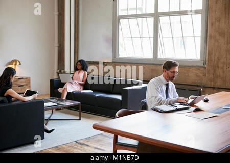 Geschäftsmann am Schreibtisch arbeiten, Kollegen auf Sofas, Seitenansicht Stockfoto