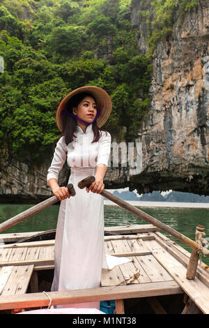 Junge Frau mit Ao Dai posieren auf einem Ruderboot in der Halong Bay, Vietnam. Die kleinen Inseln und Kalkfelsen Formationen haben sich von der UNESCO zum geschützten Bereich. Stockfoto