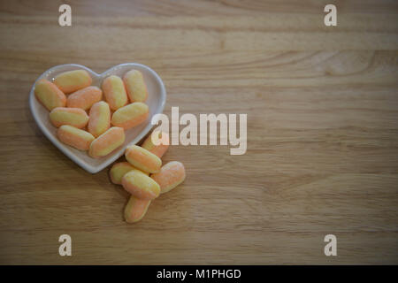 Traditionelle Spezialitäten behandelt oder Süßigkeiten in orange und gelb gestreift und mit Banane und Mango in ein weißes Herz Teller auf natürliche Holz Hintergrund aromatisiert Stockfoto