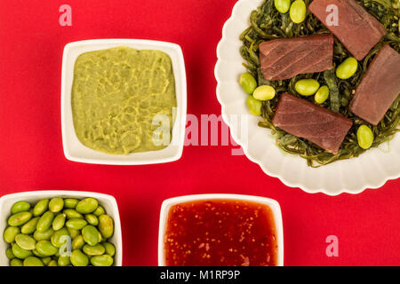 Im japanischen Stil rohen Thunfisch Sashimi auf einem Bett von eingelegtem Algen mit Edamame Bohnen auf rotem Hintergrund Stockfoto