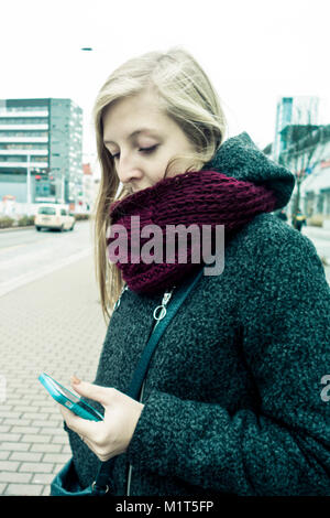 Blonde junge Frau mit Kopftuch in den Mund, in der Stadt Street, stehend und an Ihr Smartphone Stockfoto