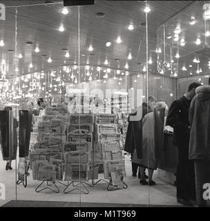 1950, historische, abends Zeit und Pariser Shopper durchsuchen Sie die Waren, die in einem gut beleuchteten glitzernde Store mit einem grossen Eingang aus Glas in der Nähe der Avenue des Champs-Elysees, mit großem Display oder Rack der französischen Tageszeitungen an der Vorderseite, Paris, Frankreich. Stockfoto