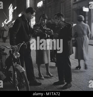1950, historische, abends mal in Paris, Frankreich, und eine Dame gibt, um etwas Geld zu einem jungen Mann mit einem Moped einen Blumenstrauß von einer Straße Käufer zu kaufen. Stockfoto