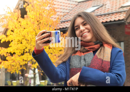 Schöne junge asiatische Frau eine selfie vor dem Hintergrund von Gebäuden und im Herbst Landschaft, hält das Telefon in der Hand, lächelt Stockfoto