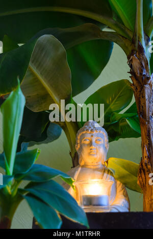 Sitzender Buddha umgeben von Bananenstauden halten Sie die brennende Kerze in der Hand Stockfoto