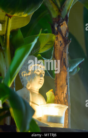 Sitzender Buddha umgeben von Bananenstauden halten Sie die brennende Kerze in der Hand Stockfoto