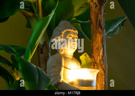 Sitzender Buddha umgeben von Bananenstauden halten Sie die brennende Kerze in der Hand Stockfoto