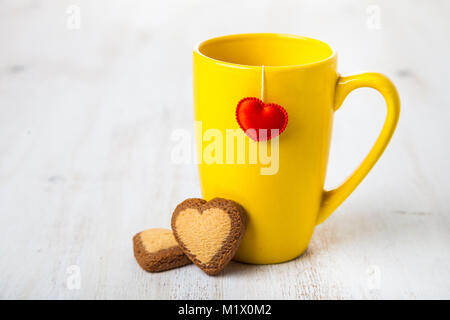 Tee in eine gelbe Tasse mit Herz und Cookies auf einer hölzernen Hintergrund für Valentinstag. Romantisches Frühstück. Grußkarte für Valentinstag Stockfoto