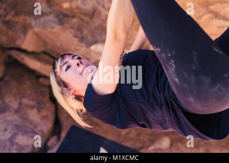 Dünne blonde kaukasische Frau mit Arm Tattoos hängt mit der Oberseite nach unten, während Sie rock auf Felsbrocken in der Wüste von Kalifornien steigt Stockfoto