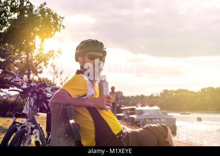 Männliche Radfahrer ruht auf einer Parkbank nach einigen Ausbildung auf seinem Fahrrad rund um die Stadt. Die Flasche in der Hand beim Stillstehen. Stockfoto