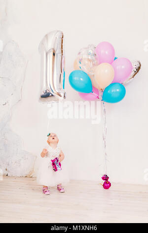 Betrifft Kinder geburtstag childs ein Jahr. Kleines Mädchen in helles Zimmer gegen weiße Wand in Kleidung und blond Holding Aufblasbare Kugel in der Form o Stockfoto