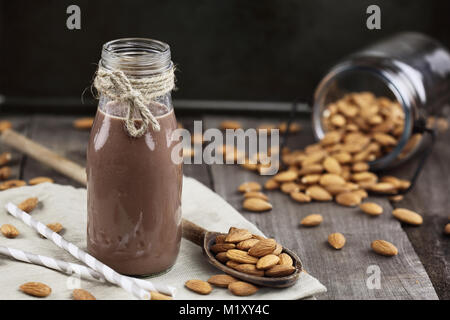 Schokolade Mandel Milch in ein Glas Flasche mit ganzen Mandeln über einen rustikalen Holztisch verschüttet. Stockfoto