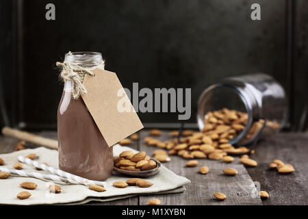 Organische Schokolade Mandel Milch mit Variablen, die in einer Glasflasche mit ganzen Mandeln über einen rustikalen Holztisch verschüttet. Stockfoto